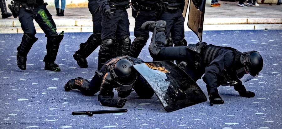 Deux CRS au sol, en armure complète. L’un est en train de se relever en s’aidant de son bouclier, la chute de l’autre n’est pas terminée : il a une jambe en l’air et des deux mains au sol. Ils sont tombés tout seuls.