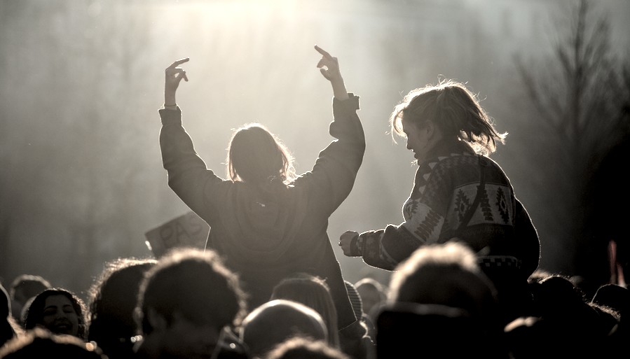 Au-dessus de la foule, deux personnes sont portées sur les épaules, leurs silhouettes se découpent à travers la fumée éclairée par le soleil couchant. L’une lève les bras et fait des doigts d’honneur.