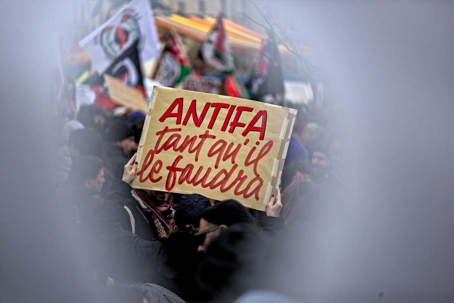 Dans la foule et ses drapeaux, une pancarte « Antifa tant qu’il le faudra ». La photo est prise au travers d’un trou dans un tissu, qui remplit de blanc le reste de l’image.